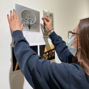 A student wearing a mask, hanging photographs on a classroom wall.